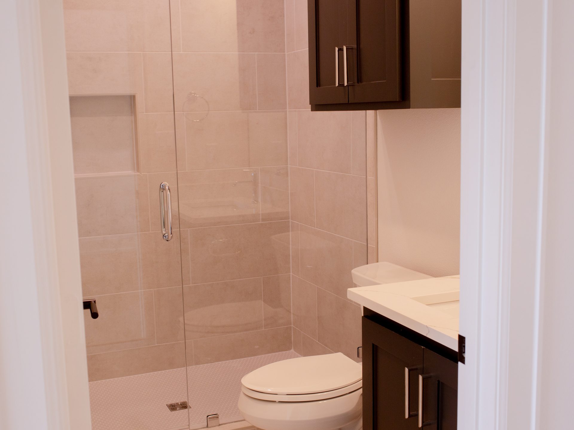 Walk-in shower with frameless glass door, large-format tan tile walls, and dark vanity cabinetry in the foreground.