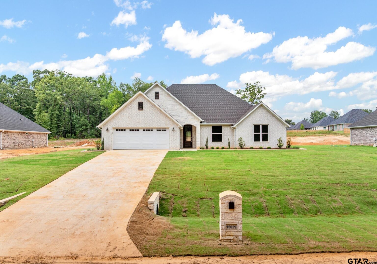 White brick single-story Nicholas Humphrey home at 15029 County Road 1198, with gray roof, attached two-car garage, and a wide front lawn backed by woods.