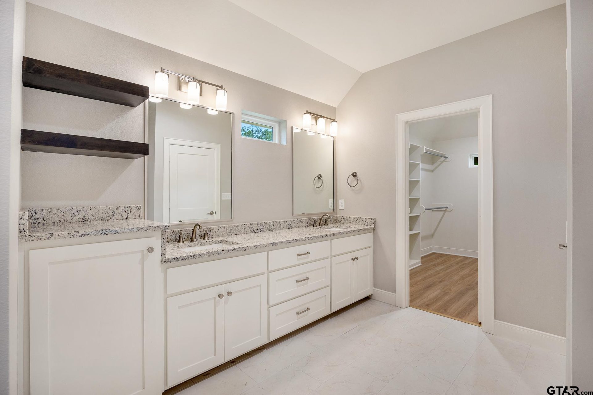 Primary bathroom with a double granite vanity, floating dark wood shelves, and a view into the walk-in closet beyond.