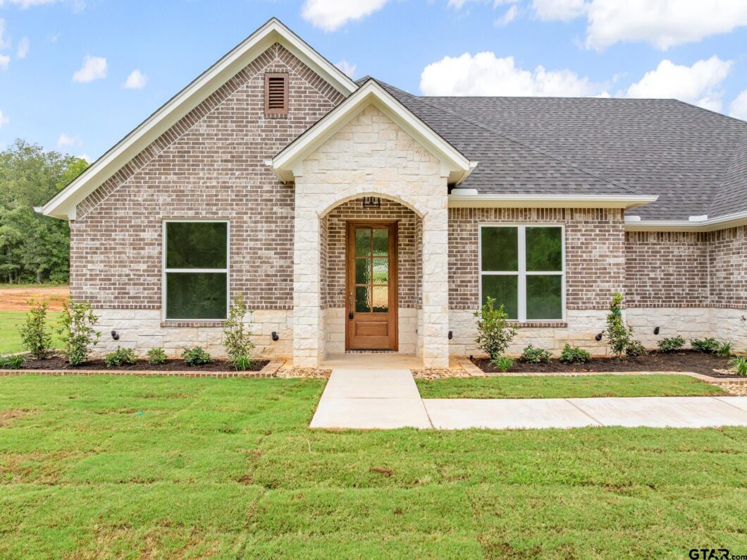 Front entry of 15051 County Road 1198 with mixed brick and stone, an arched wood door, and neat landscape beds.