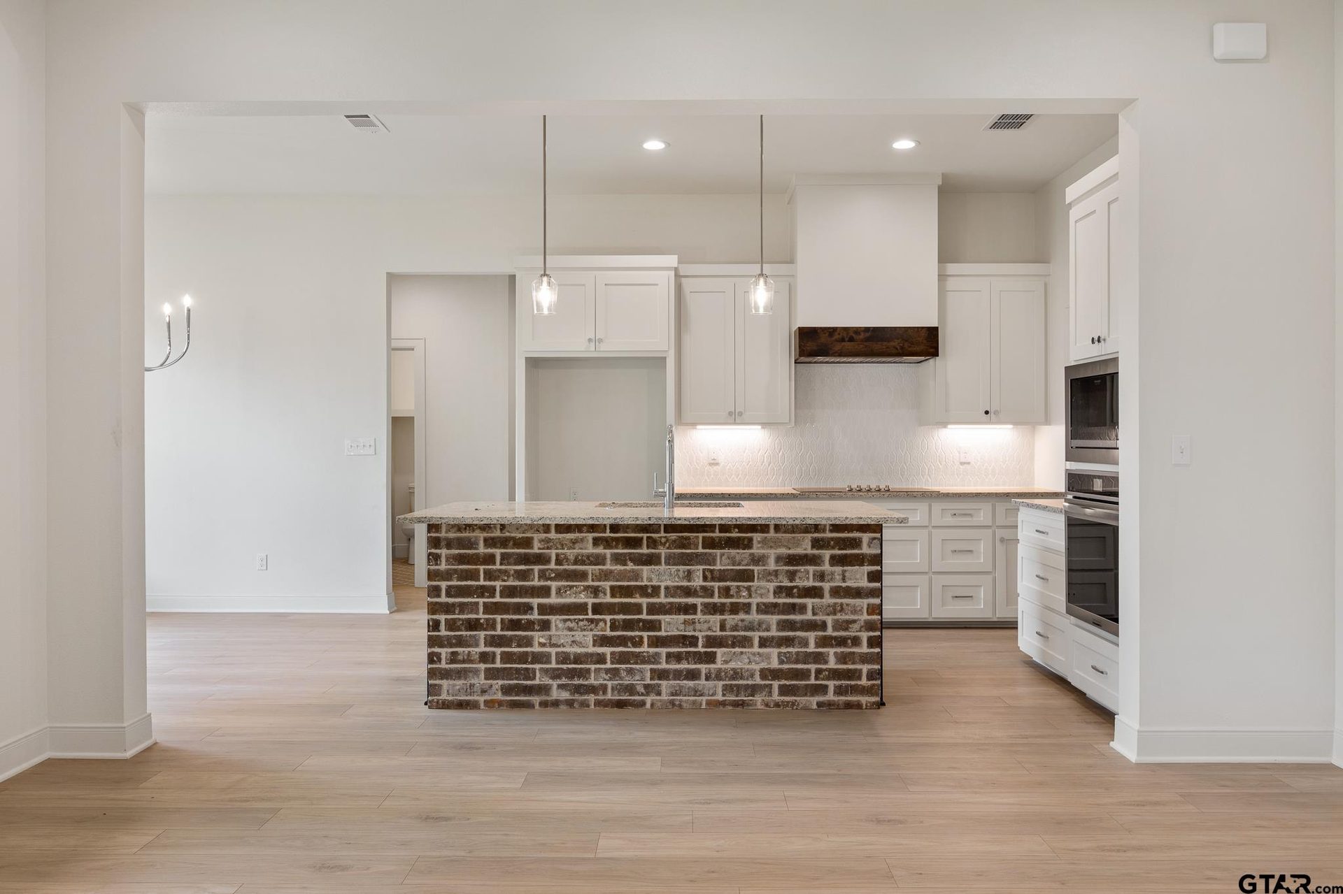 Kitchen with a reclaimed-brick waterfall island, white cabinetry, a wood range hood, and open dining beyond on light oak floors.