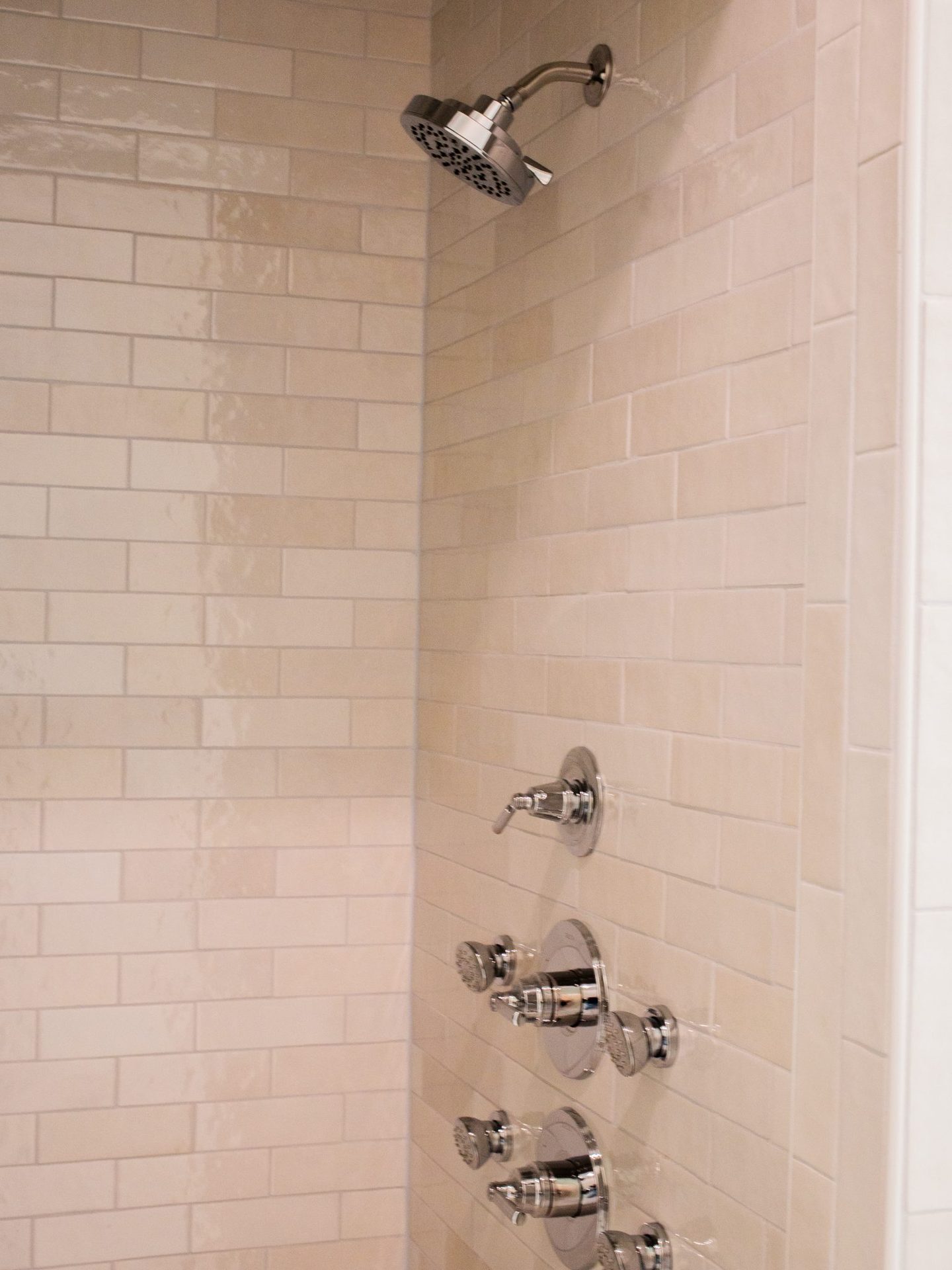 Shower detail with handheld and overhead shower heads and chrome fixtures on a cream subway-tile wall.